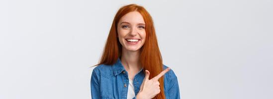 Close-up portrait outgoing good-looking cheerful redhead teenage girl in casual outfit, laughing and talking, discuss recent event, pointing finger left at banner, recommend advertisement photo