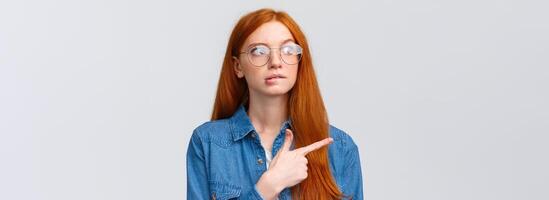 Unsure smart and cute redhead teenager in glasses, female student biting lip curious looking and pointing left, thinking, deciding what pick, making choice, standing over white background photo