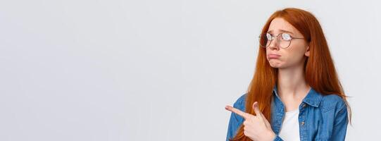 Close-up portrait gloomy and sad pouting cute redhead girl with lovely blue eyes, wear glasses, sulking and sobbing sad, pointing looking upper left corner with regret or jealousy, want something photo