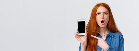 Close-up portrait angry and pissed-off redhead girl showing photos from social media ex-boyfriend complaining to friend, pointing finger smartphone display and frowning upset, white background