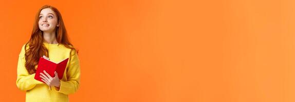 Dreamy, inspirational cute redhead female student writing daily tasks, to-do list in red lovely planner, look upper left corner thoughtful as smiling, getting inspiration to write novel, orange wall photo