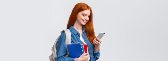 Modern lifestyle, technology and education concept. Cute, lovely redhead female student heading to campus after classes in college, holding backpack and notebooks, chatting on smartphone photo