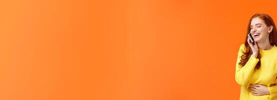 Communication, technology and people concept. Cheerful pretty redhead hipster girl talking on phone and laughing carefree, touching belly close eyes, smartphone near ear, orange background photo