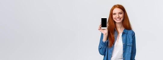 Cheerful friendly redhead woman checking water app, stay hydrated with smartphone application, showing mobile screen and smiling delighted, standing white background in casual clothes photo