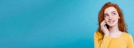 Lifestyle and Technology concept - Portrait of cheerful happy ginger red hair girl with joyful and exciting talking with friend by mobile phone. Isolated on Blue Pastel Background. Copy space photo