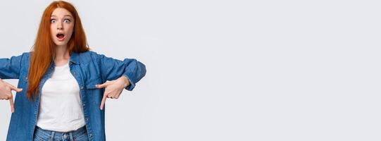 Amazing, have you heard already. Astonished, speechless and impressed redhead woman in denim shirt, drop jaw, gasping stare camera fascinated, say wow, pointing fingers down photo