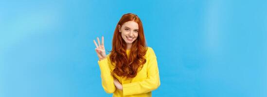 Gorgeous young redhead girl 20s in yellow sweater, smiling carefree making order, reservetion for three people, showing number third with fingers, standing blue background photo