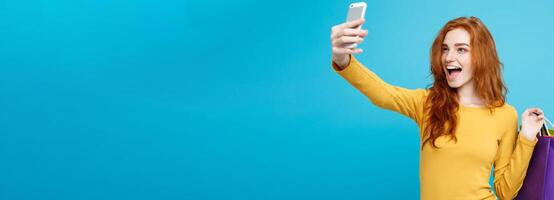 Shopping Concept - Close up Portrait young beautiful attractive redhair girl smiling looking at camera with white shopping bag and selfie. Blue Pastel Background. Copy space photo