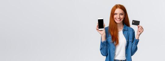 Waist-up portrait confident female with red long hair, blue eyes, holding credit card and showing smartphone display, easy banking service, payment for bills internet, smiling satisfied photo
