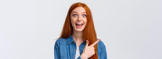 Close-up portrait excited happy ginger girl, foxy hair and blue eyes, smiling upbeat with amazement and joy, pointing finger left, attend amusing cool party, telling about it, white background photo