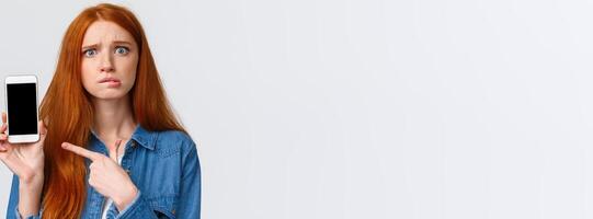 Worried, insecure and alarmed cute redhead girl asking for advice having problems messaging with stranger on dating app, dont know what answer standing nervous pointing finger at smartphone display photo