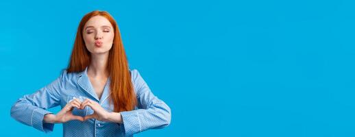 Kiss for goodnight. Charming pretty redhead female in pyjama folding lips and close eyes give mwah, showing heart gesture over chest, express affection or tender feelings, blue background photo