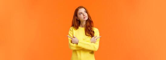 Pensive girl trying make decision, thinking, looking upper left corner thoughtful, pondering what buy for holidays, pointing sideways left and right, looking up dreamy, orange background photo