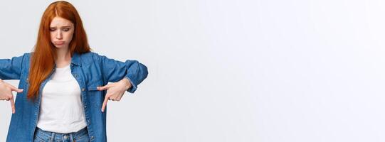 Upset, gloomy timid redhead teenage girl express pity or jealousy, regret something broken, sobbing, pouting sad and looking pointing down distressed, standing unhappy over white background photo