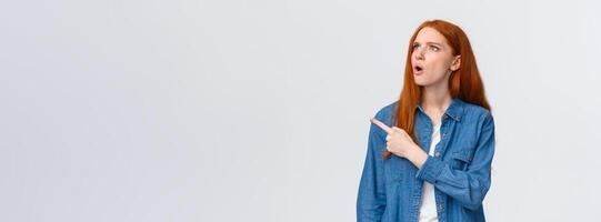 Perplexed and indecisive redhead cute girl cant understand wtf is that, pointing looking upper left corner with confused expression, dont know and asking question, standing white background photo