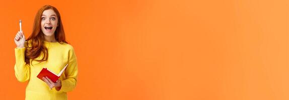 Inspired and dreamy, smart redhead female student got excellent idea for her book, write in notebook plans and to-do list, raising pen in eureka gesture and smiling astonished, orange background photo