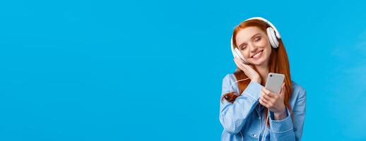 Tenderness, women and lifestyle concept. Cheerful tender redhead female in pyjama, tilting head enjoying listening music, holding smartphone and smiling as touching earphones from delight photo