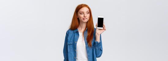 Curious and daring sassy redhead girl tempting buy something in online store, have interesting plan, smirk and peek at smartphone screen with pleased expression, standing intrigued white background photo