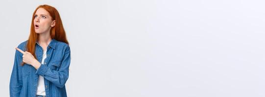 Perplexed and indecisive redhead cute girl cant understand wtf is that, pointing looking upper left corner with confused expression, dont know and asking question, standing white background photo