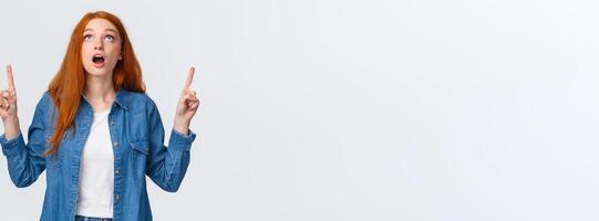 Waist-up portrait amazed and impressed, curious redhead girl checking out awesome game in store, drop jaw, looking and pointing up speechless, gasping amused, stand white background photo