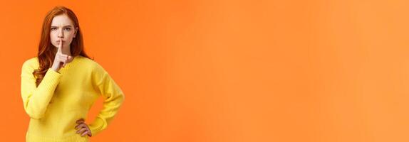 Serious and mad elder sister say keep silent, forbid talking during class, shushing angry with index finger pressed mouth, hold hand on waist in dissatisfied pose, standing orange background photo