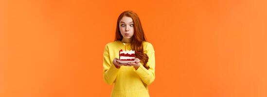 Funny and silly cute redhead b-day girl squinting eyes making goofy expression, pouting as holding birthday cake and blowing-out candle to make wish, having fun at party celebrating, orange wall photo
