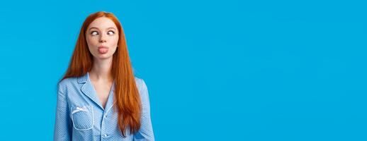 Be funny and silly. Redhead carefree and enthusiastic caucasian girl squinting, making goofy expression, showing tongue, standing blue background in nightwear, fool around photo