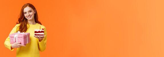 Cute redhead girl coming party with wrapped gift and piece of delicious b-day cake, asking friend blow-out candle and make with celebrating girlfriends birthday, smiling over orange background photo