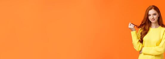 Gorgeous flirty and cute young modern redhead cheeky girl, rolling curl on hair and glancing camera curious, with interest and coquettish flirting smile, standing orange background intrigued photo