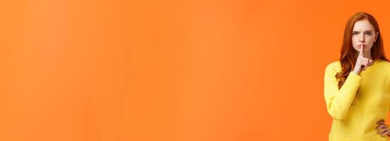 Serious and mad elder sister say keep silent, forbid talking during class, shushing angry with index finger pressed mouth, hold hand on waist in dissatisfied pose, standing orange background photo