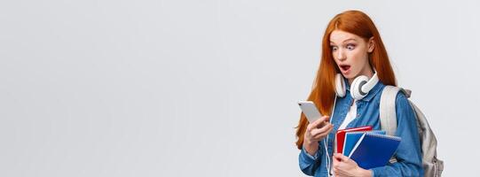 Shocked and astonished, gasping speechless redhead female student with backpack and notebooks, staring at smartphone screen shook open mouth and pop eyes mobile display photo