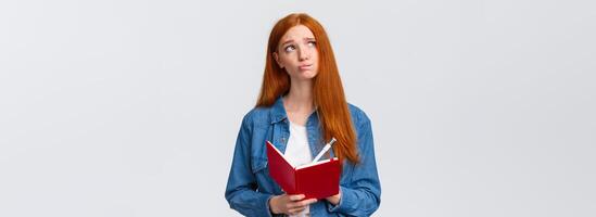 Uneasy and gloomy, indecisive cute redhead girl writing down her feelings in red notebook, looking nervous and troubled up thinking, holding pen, pondering something while sharing thoughts with paper photo
