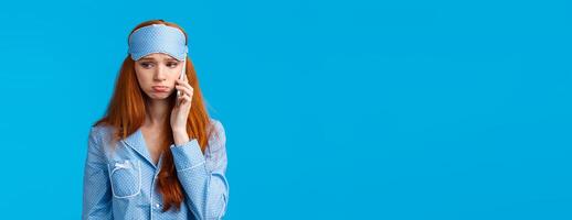 Sulky and gloomy upset cute silly redhead girl calling girlfriend talking about problems, had bad dream, holding smartphone near ear, having phone call in nightwear and sleep mask photo
