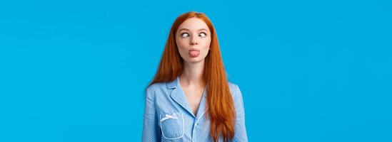 Be funny and silly. Redhead carefree and enthusiastic caucasian girl squinting, making goofy expression, showing tongue, standing blue background in nightwear, fool around photo