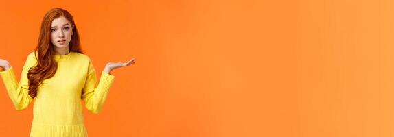 Perplexed cute redhead girl, shrugging with raised arms and looking confused, dont know answer, cant help, feeling confused or indecisive, cannot understand, standing orange background photo