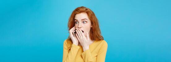 Lifestyle concept - Portrait of ginger red hair girl with shocking and stressful expression while talking with friend by mobile phone. Isolated on Blue Pastel Background. Copy space. photo