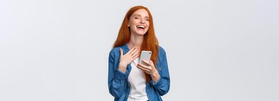 Waist-up portrait carefree and joyful, alluring redhead caucasian girl receive funny meme message, holding smartphone, laughing and close eyes dreamy, standing white background photo