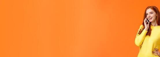 Outgoing pretty redhead girl having conversation on phone, calling friend, making arrangements, woman booking hotel or trip using mobile, speaking via smartphone, standing orange background photo