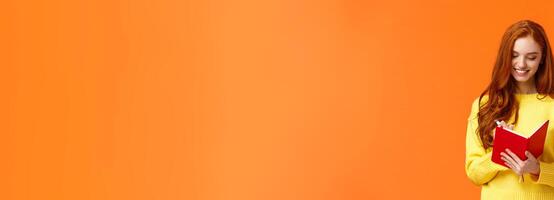 Waist-up portrait attractive redhead girl 20s write in her diary, fill-in to-do list or personal schedule in new notebook, smiling as having everything planned, standing orange background carefree photo