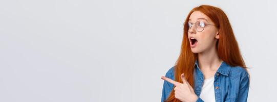 Cool take a look. Close-up portrait fascinated and excited cute redhead woman in glasses checking out awesome banner, pointing and looking left with opened mouth, say wow, white background photo