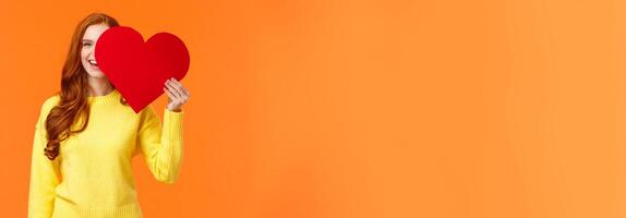 Holidays, romance concept. Cheerful girl showing affection, give heart to girlfriend, hiding half face behind valentines card, smiling joyfully, prepare romantic gesture for date, orange background photo