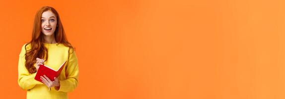Inspired and upbeat, dreamy fascinated redhead girl writing down interesting lecture, smiling and gazing with admiration or enthusiasm, fill-in notebook schedule, standing orange background photo