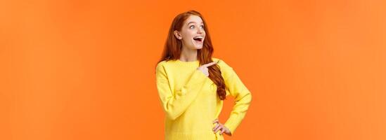Look at that, wow. Impressed and fascinated, astonished cute redhead woman stare and pointing left with excited, happy smile, shoppaholic adore winter holiday sales, express interest and admiration photo