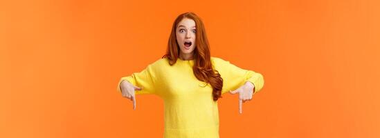 Take look quick. Amazed and fascinated cute ginger girl, redhead student in yellow sweater, gasping open mouth from surprise and wonder, pointing fingers down, look impressed, orange background photo