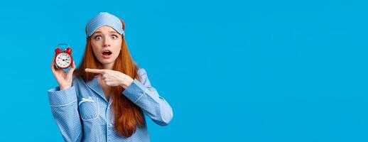 Shocked and concerned, worried cute redhead girl pointing at alarm clock with frustrated, nervous expression, being late, dont know how set up right time, standing blue background in nightwear photo