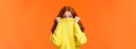 Surprised and astonished cute curious redhead female pulling sweater collar on face and popping eyes at camera with amazed expression, found something interesting, orange background photo