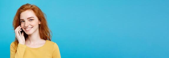 Lifestyle and Technology concept - Portrait of cheerful happy ginger red hair girl with joyful and exciting talking with friend by mobile phone. Isolated on Blue Pastel Background. Copy space. photo