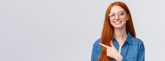 Close-up portrait cute cheerful redhead, foxy girl in glasses, showing place to click website, follow or subscribe, pointing finger left and smiling pleasant, with friendly attitude, white background photo