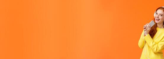 Pretty redhead caucasian woman enjoy listening music, playing karaoke app on smartphone with headphones on, looking camera joyfully, hold mobile phone like microphone, orange background photo