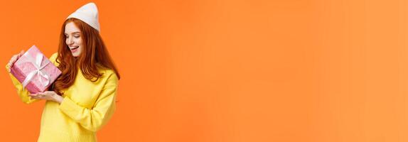 Girl trying figure-out whats inside gift box shaking it and looking intrigued. Excited happy redhead woman like winter holidays and receiving present, standing amused orange background photo